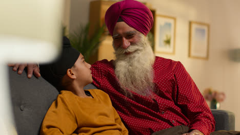 Smiling-Sikh-Grandfather-And-Grandson-Wearing-Turbans-Sitting-On-Sofa-At-Home-Talking-Together